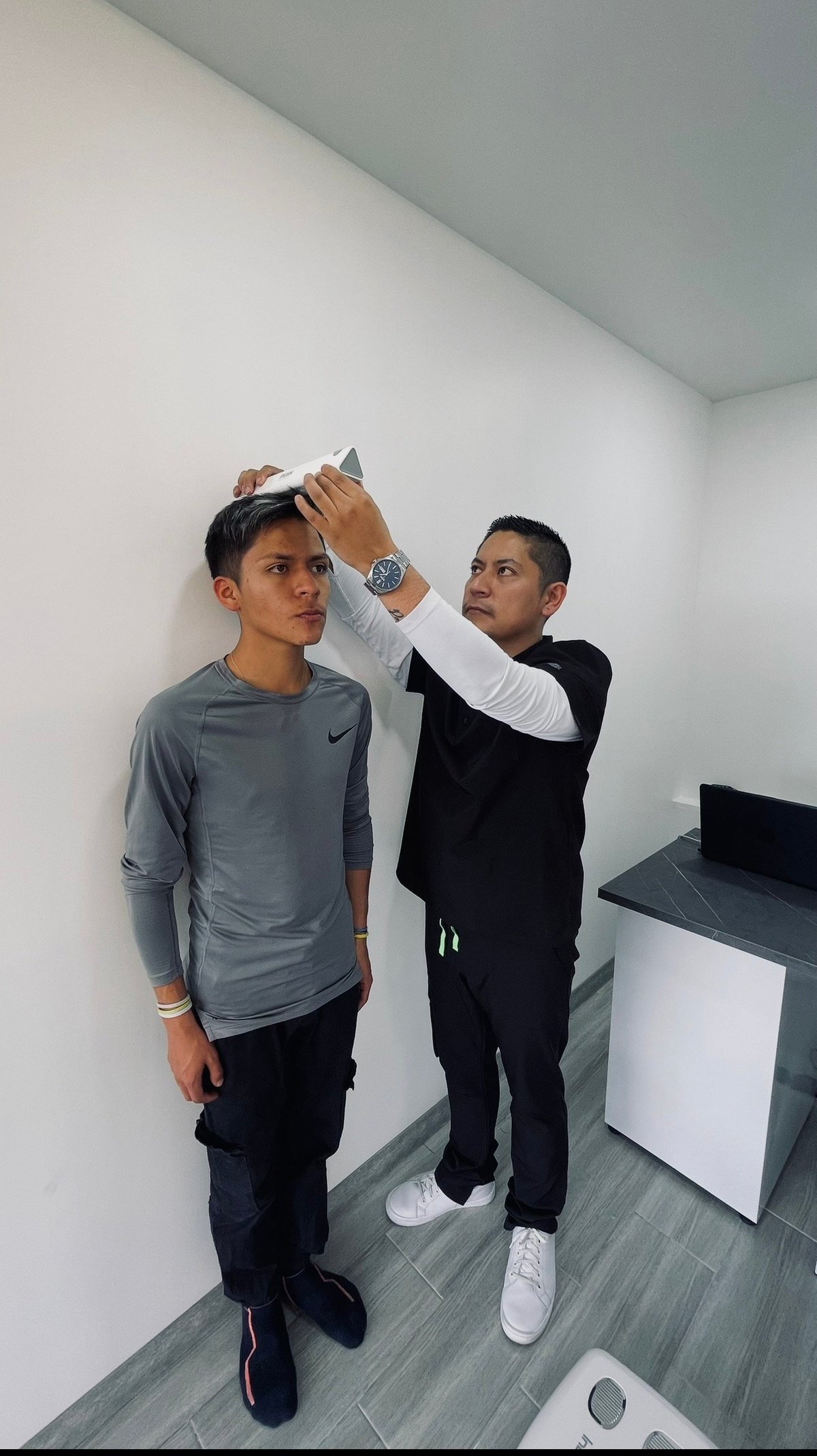 Dos jóvenes en una habitación blanca; uno con camisa Nike gris midiendo la estatura del otro contra la pared