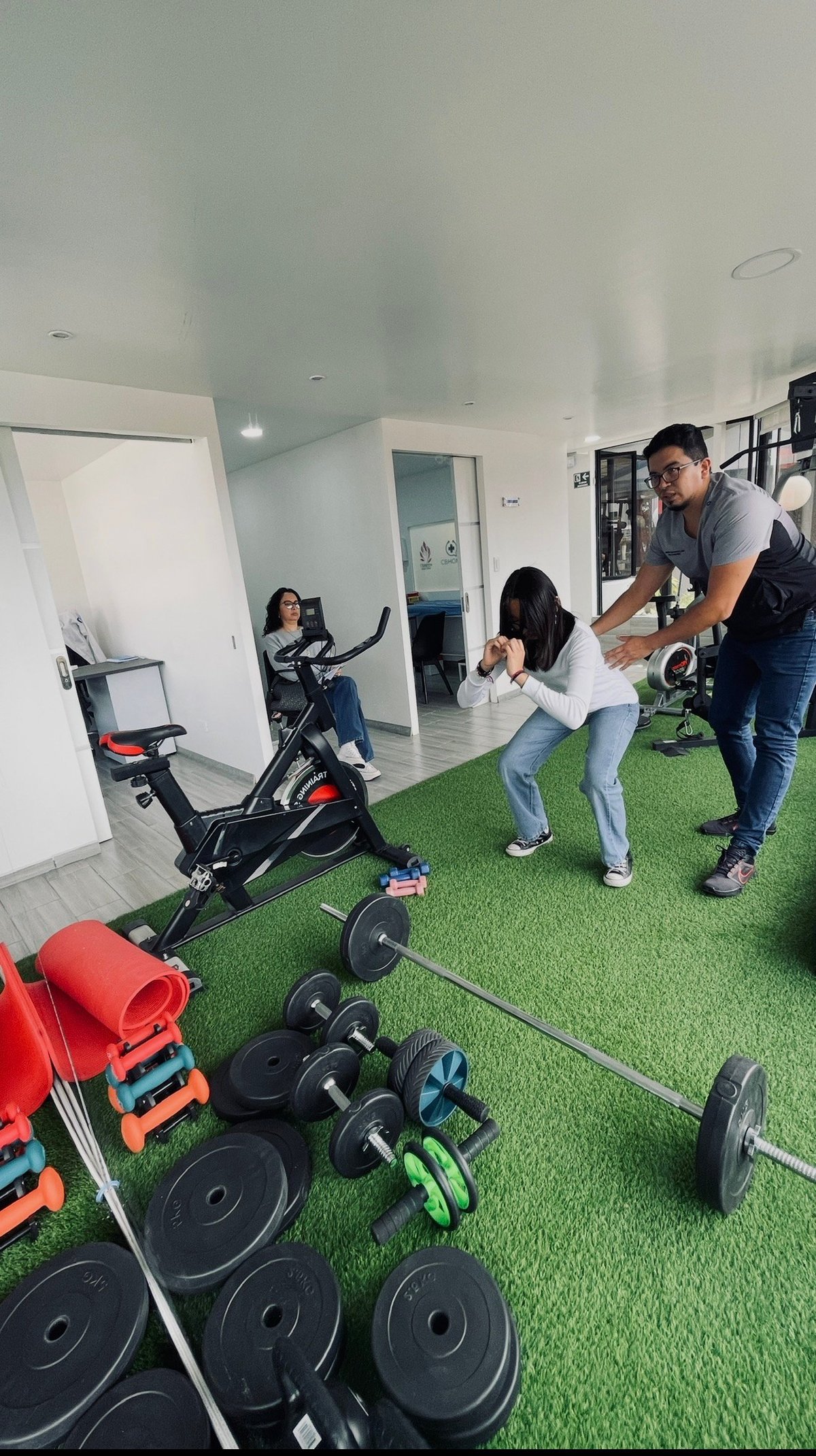 Indoor gym with trainer instructing two women exercising on artificial turf, surrounded by weights and stationary bike equipment