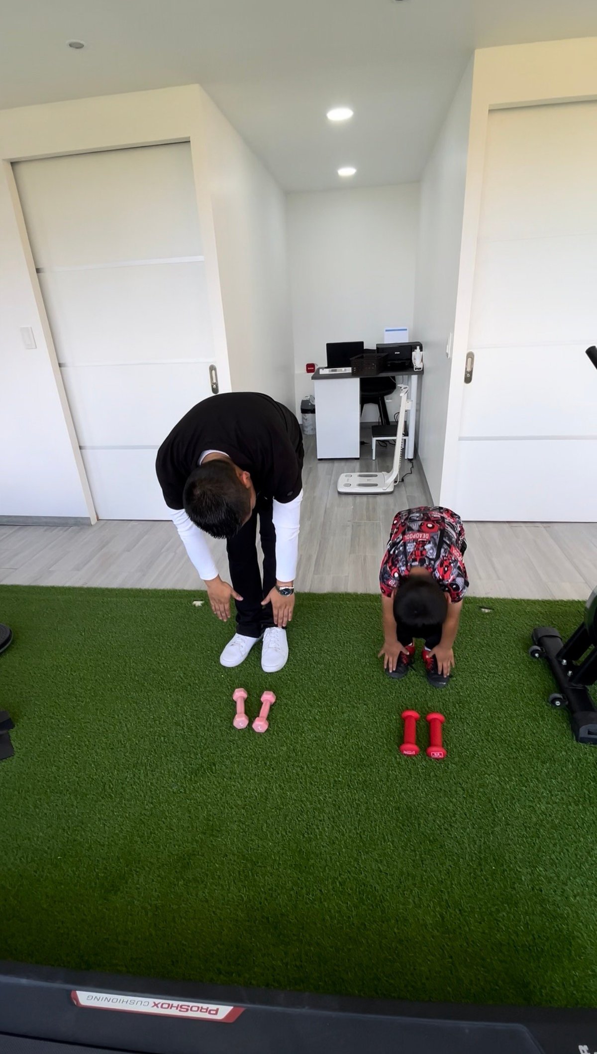 Two people exercising indoors on artificial turf, bending forward with dumbbells placed on the ground between them