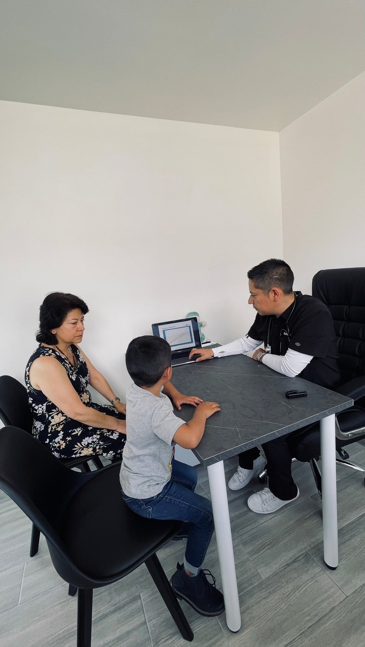 Three people at a table during a consultation or meeting, with a laptop visible and white office walls