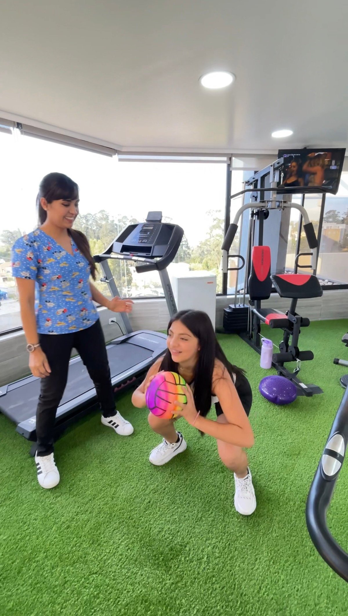 Two women exercising in a modern gym with equipment including treadmill, weights, and cardio machines on artificial turf flooring